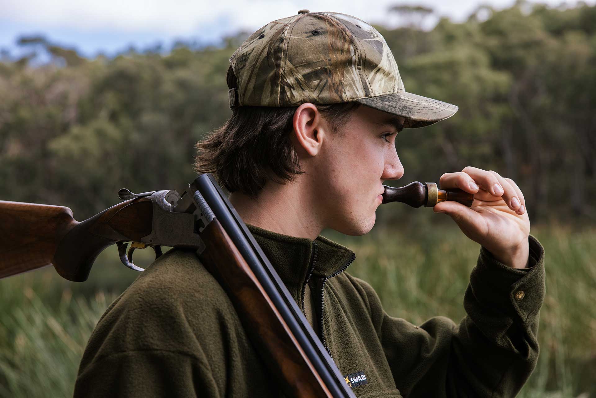 A hunter wearing camouflage gear and with a gun over his shoulder blows into a duck caller.